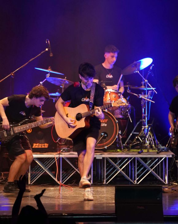 Concerto live con studenti della Scuola e Accademia di Musica Cluster: chitarrista acustico, bassista e batterista si esibiscono su palco illuminato, in un evento musicale scolastico